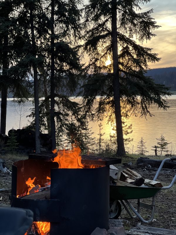 Campfire burning in a metal fire ring beside Leighton Lake at sunset, with firewood in a wheelbarrow nearby